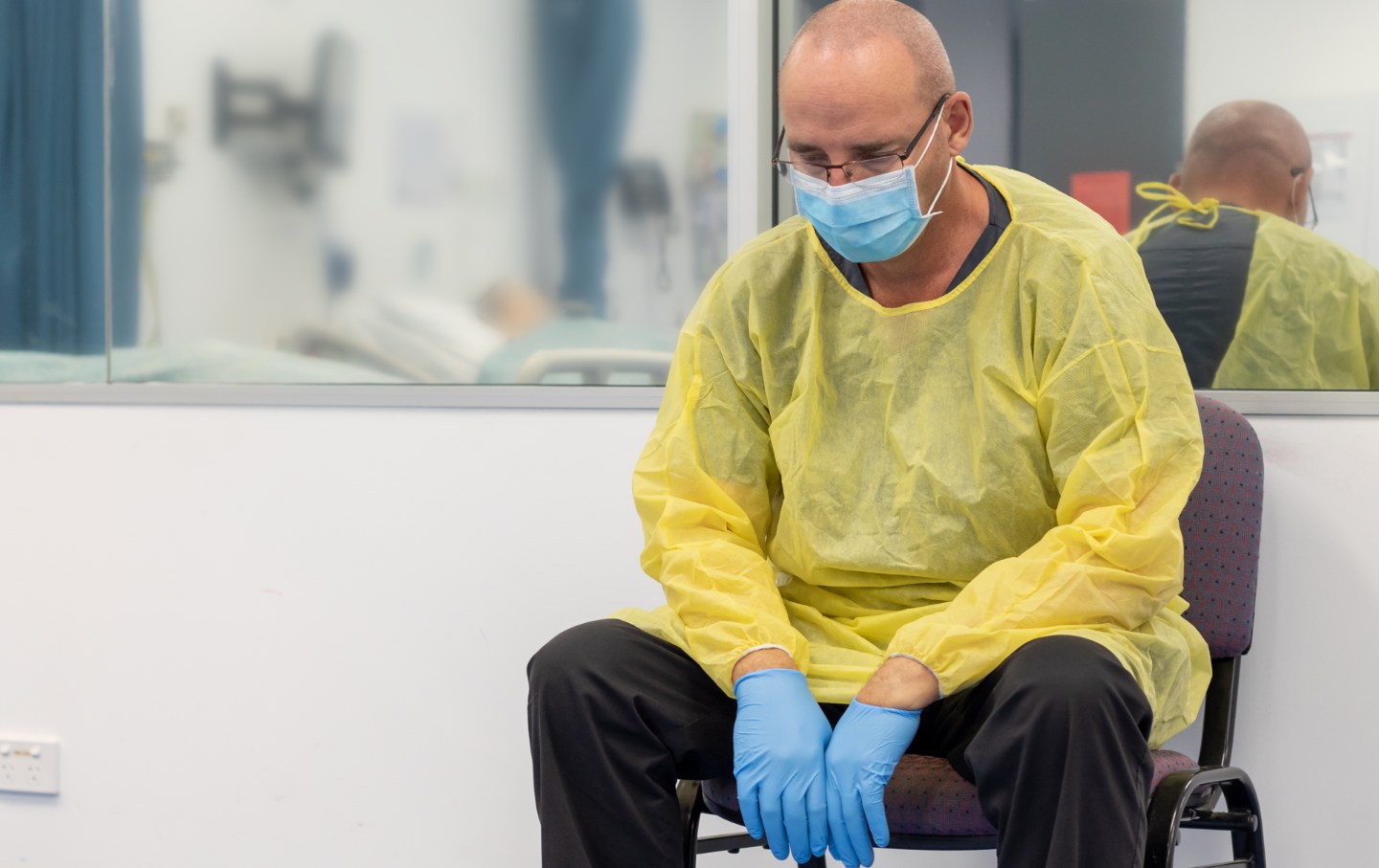 Close up of a Male Health care worker wearing protective equipment, mask, gown and gloves, sitting in a chair beside a closed hospital room looking exhausted.
