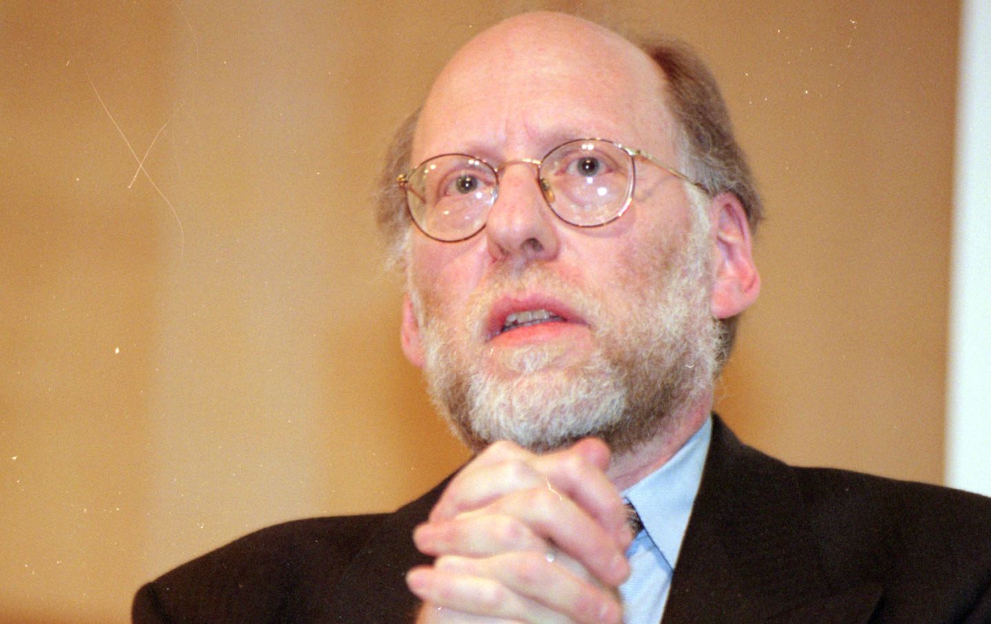 A man with a beard and glasses looks into the distance. His hands are clasped below his chin.