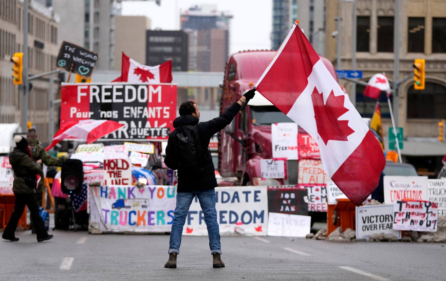 A man with his back to the camera waves a Canadian flag in front of a protest