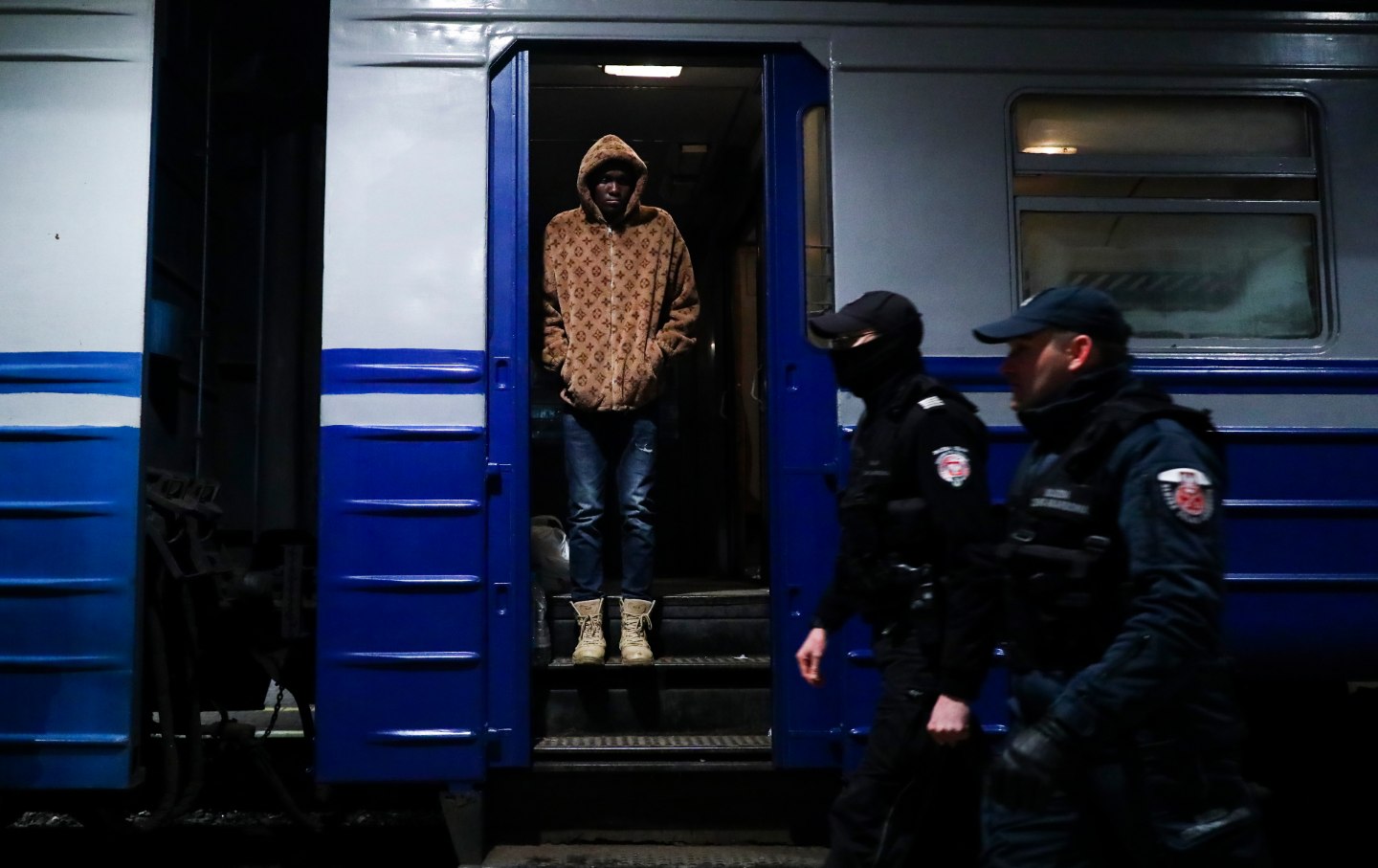 A dark skinned man in a printed coat, jeans and boots stands on a train platform as two men in uniform walk by