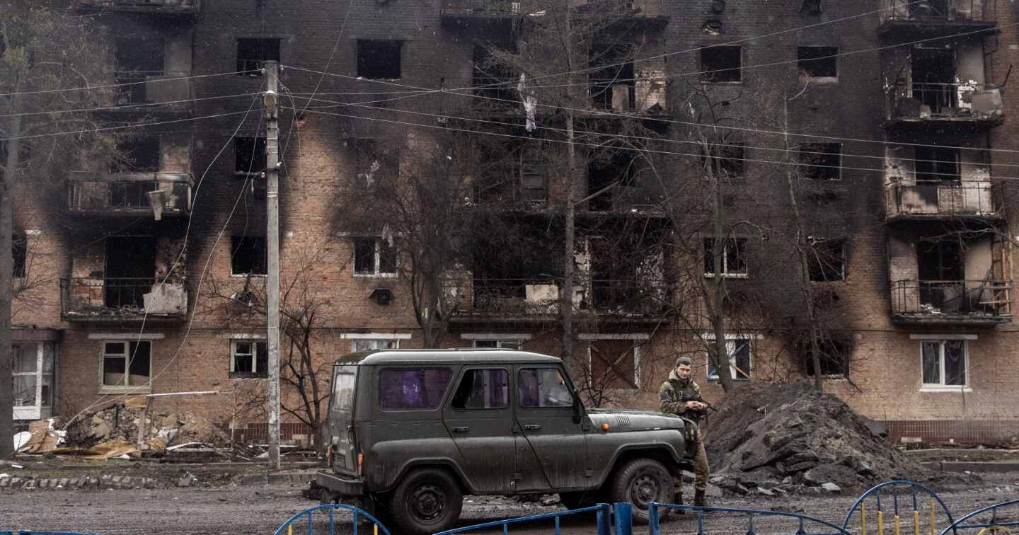 A car sits in front of a burned apartment building and pile of rubble. A soldier stands in front of the car.