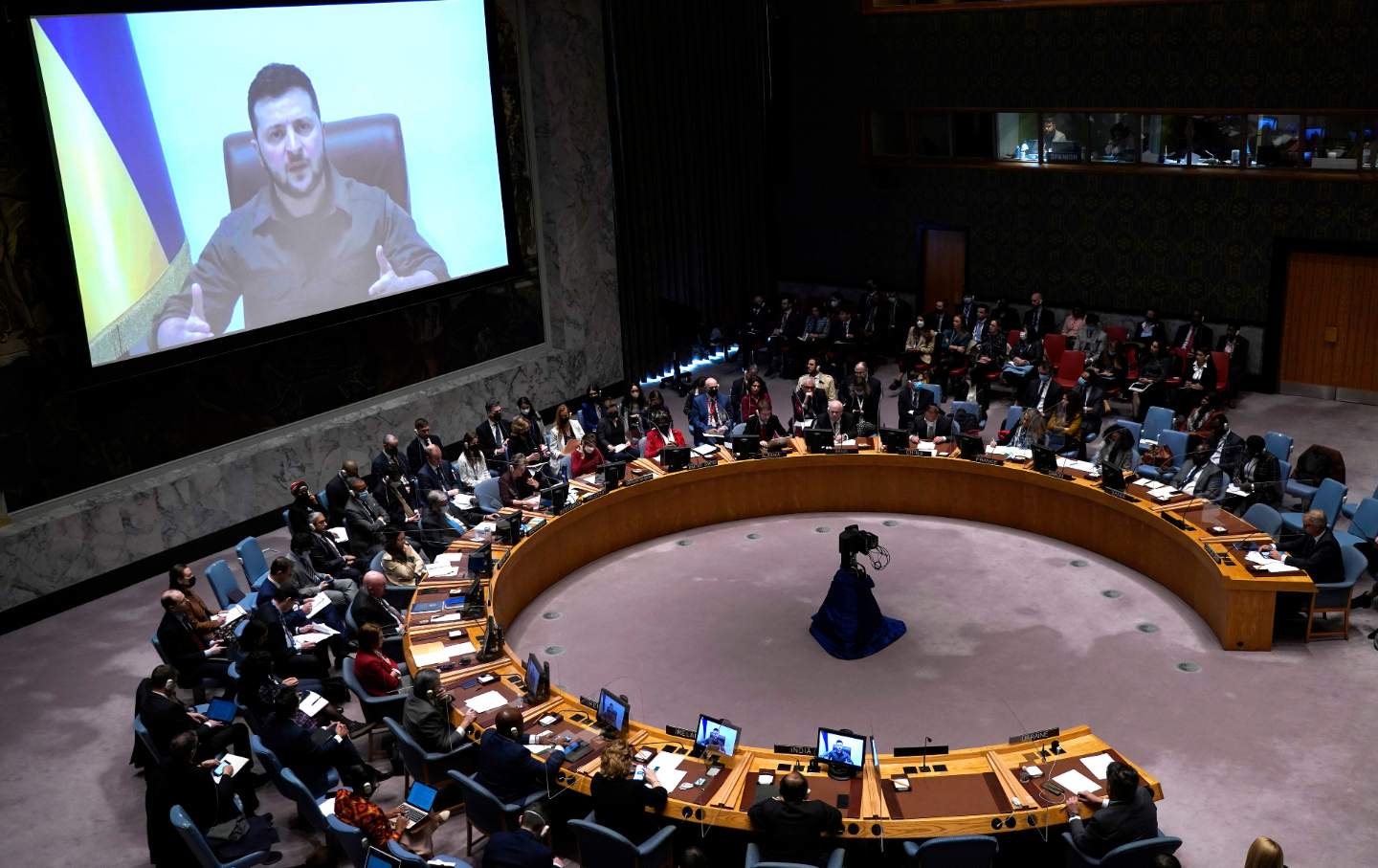 People seated around a horseshoe shaped table watch an on-screen address from Ukraine's president