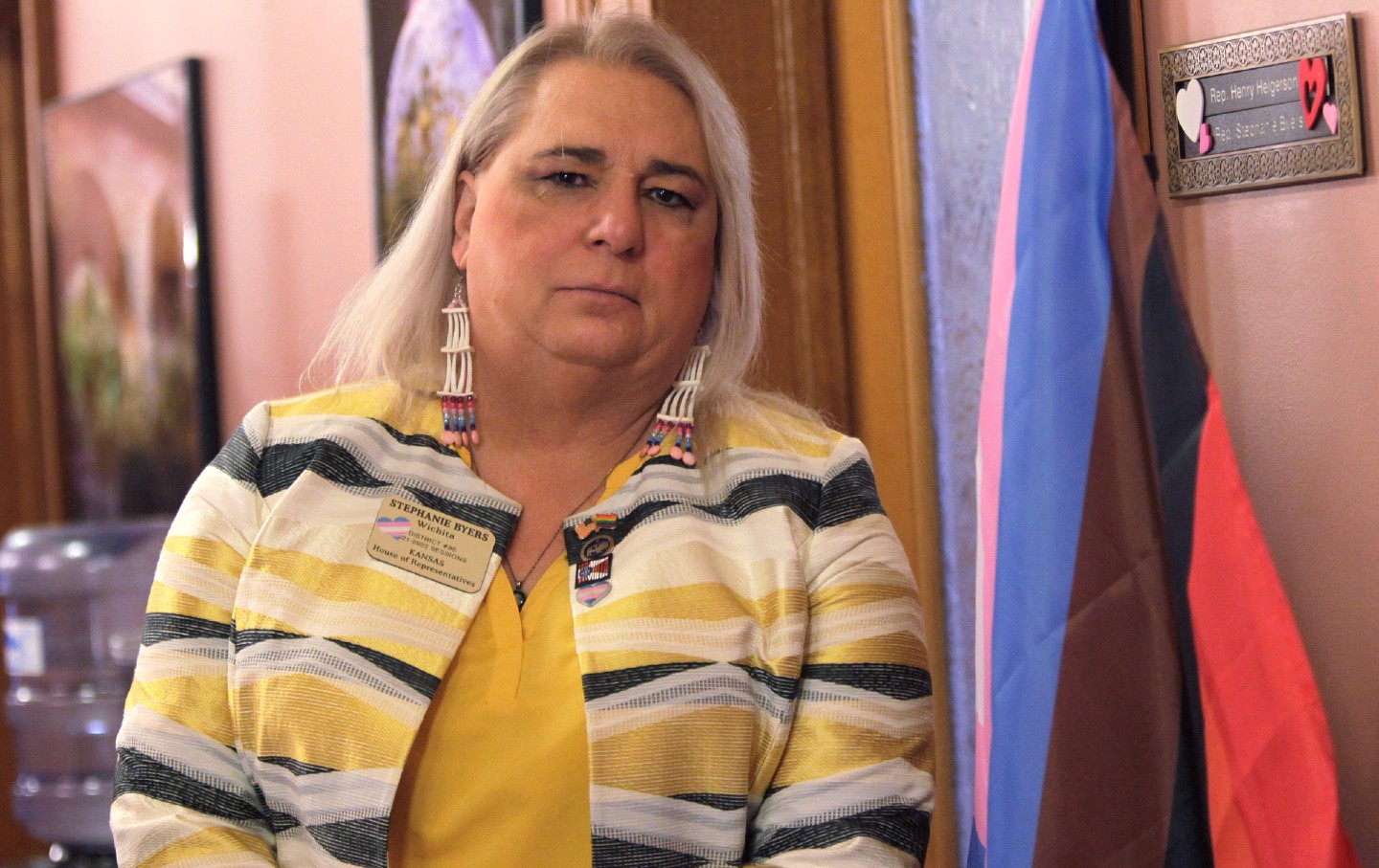 Stephanie Byers stands outside her office, wearing beaded earrings and a nametag decorated with a trans flag sticker.