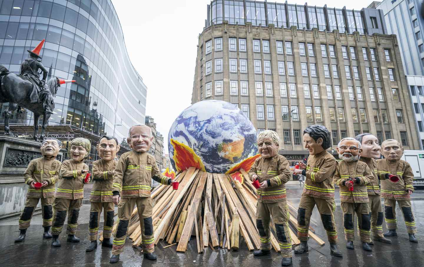 People wearing fire suits and masks of world leaders stand around a sculpture of the Earth on a pyre.