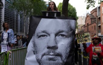 A woman gives a speech at a lectern draped with a picture of Julian Assange.