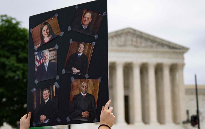 Hands hold up a poster with pictures of the six conservative Supreme Court justices with their eyes crossed out.