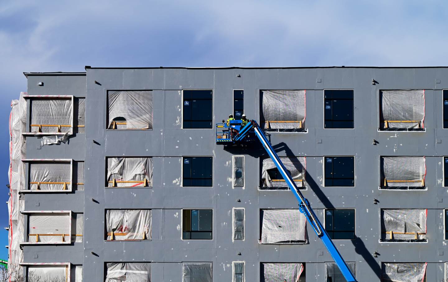 Workers in yellow vests and hard hats on a blue crane in front of a gray building