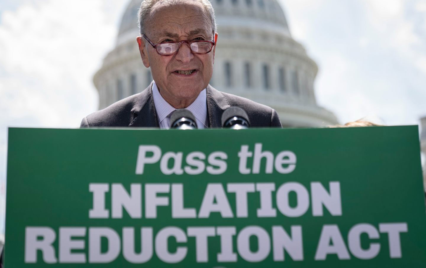 Senator Chuck Schumer speaks outside the U.S. Capitol about the Inflation Reduction Act. He stands in front of the Capitol building and in front of a green sign that reads 