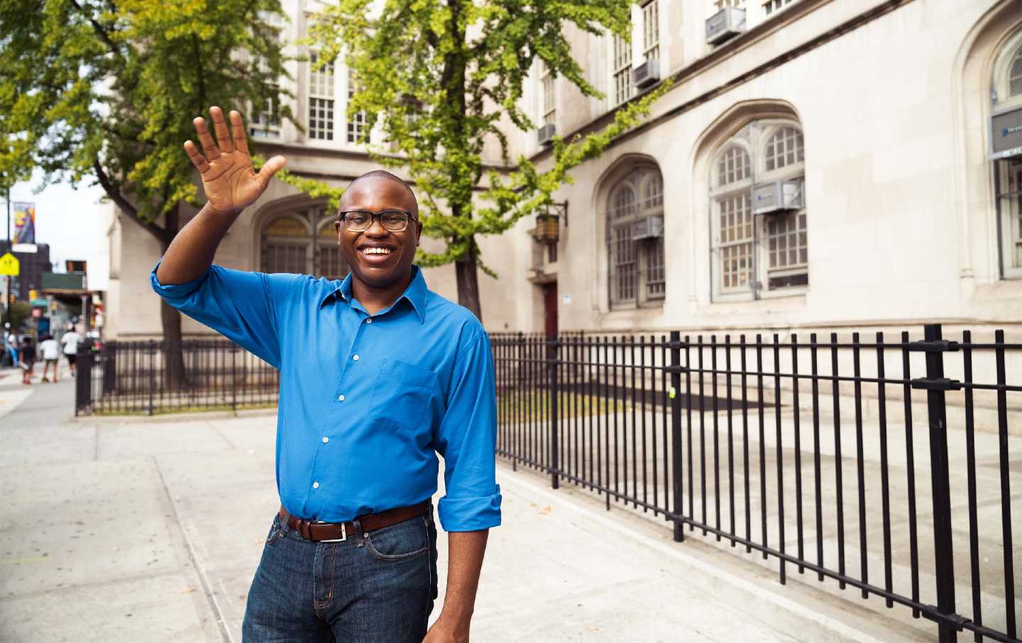 David Alexis smiles and waves to the camera, wearing a blue shirt and jeans outside a Brooklyn high school.