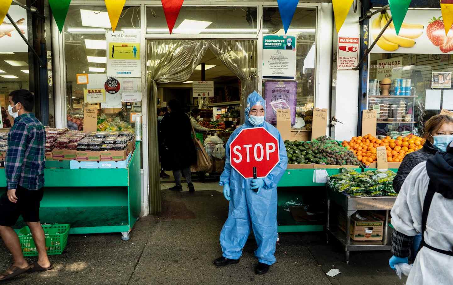 Outside a supermarket, a masked essential worker wearing a blue hazmat suit holds a stop sign.