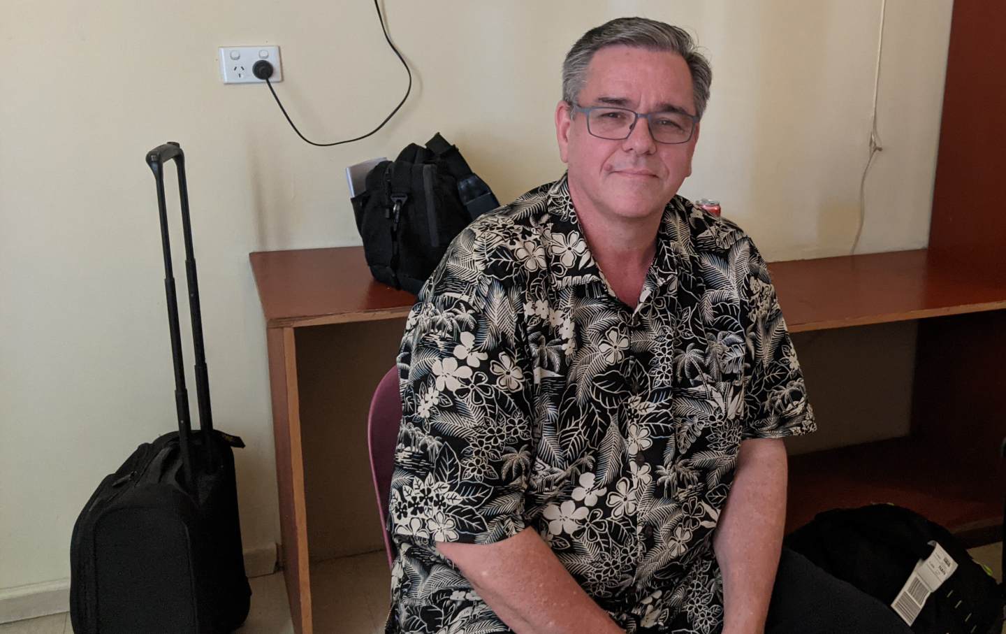 David Lambourne sits in a tropical shirt alongside some luggage.