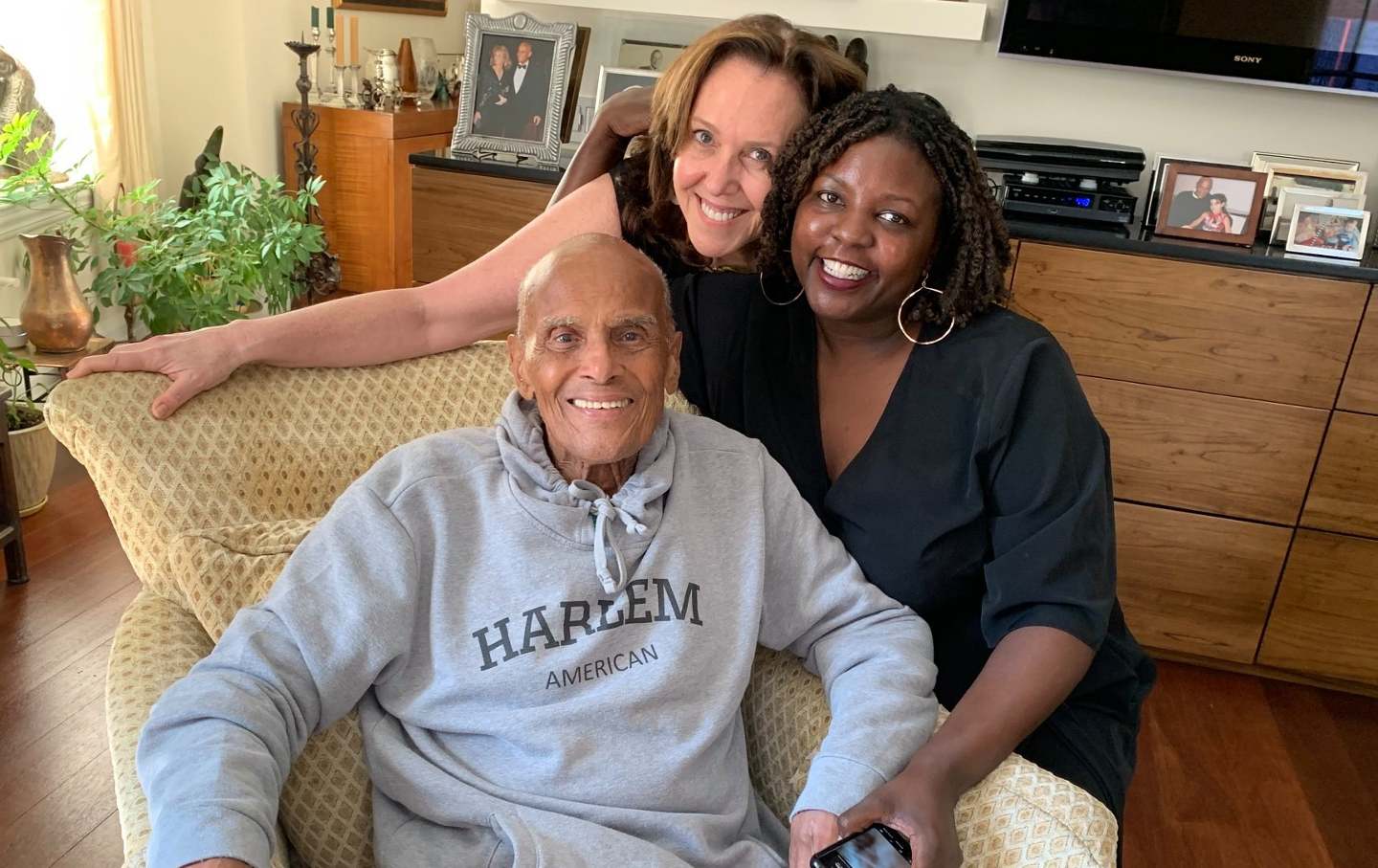 Harry Belafonte, seated in an armchair, with Joan Walsh and Yoruba Richen standing behind him. All three are smiling.