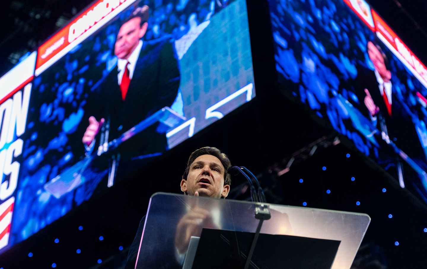 Florida Governor Ron DeSantis at a podium, seen from below, with his image on a large TV screen above him.