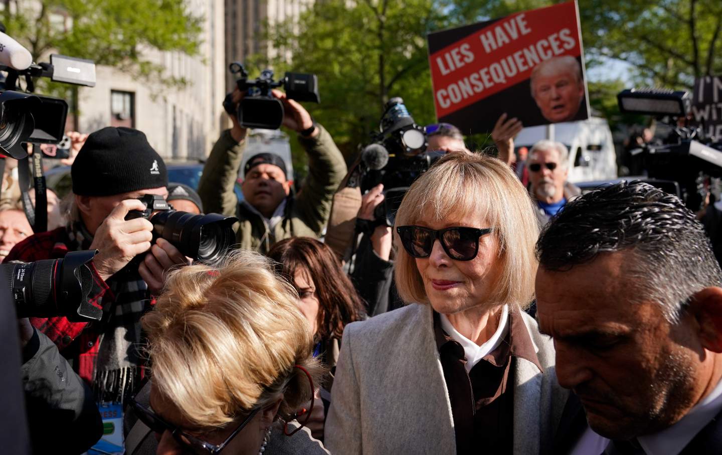 E. Jean Carroll outside Manhattan federal court house surrounded by a scrum of people, one holding a sign saying 