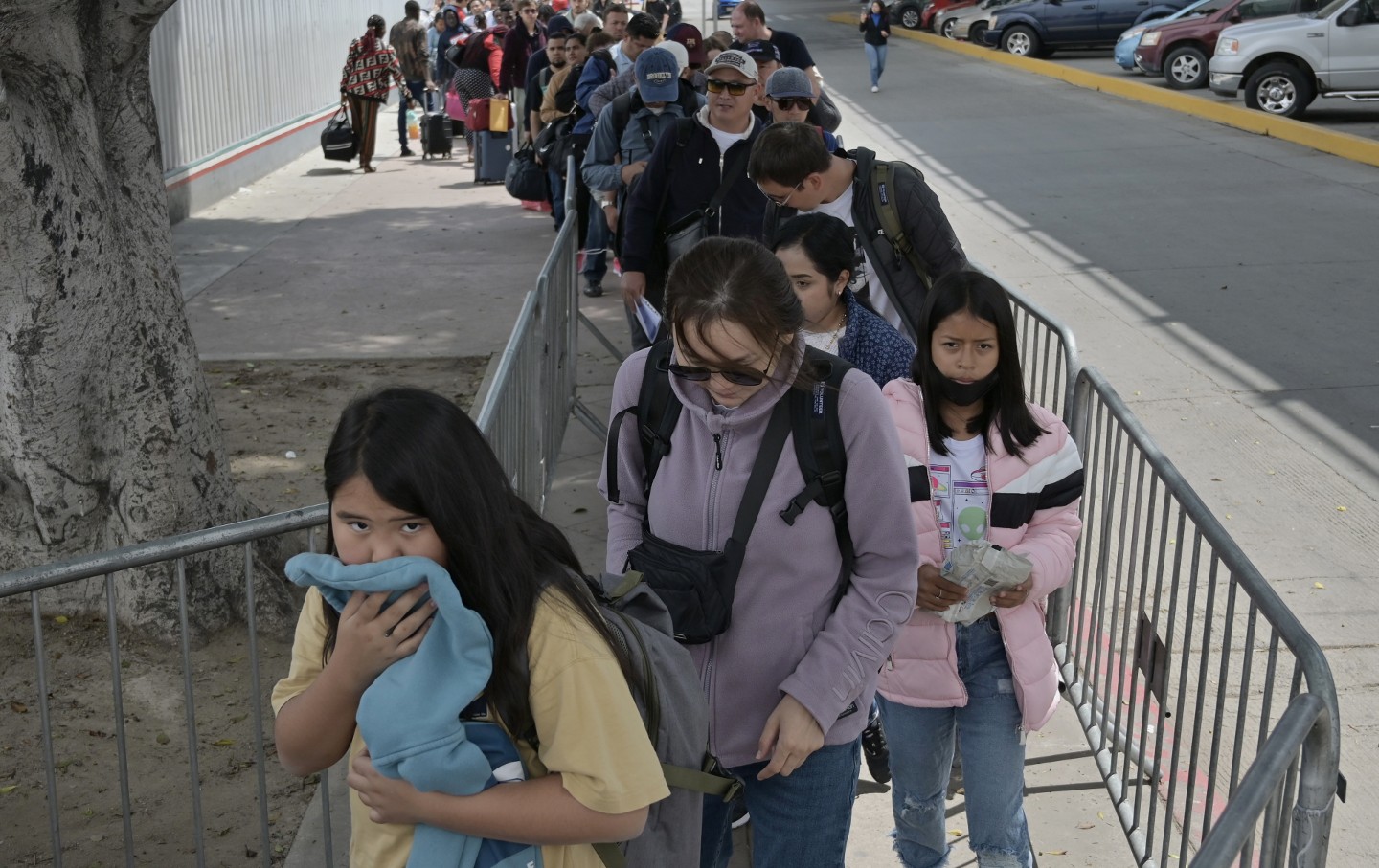 A line of migrants carrying their possession stand between metal fencing.