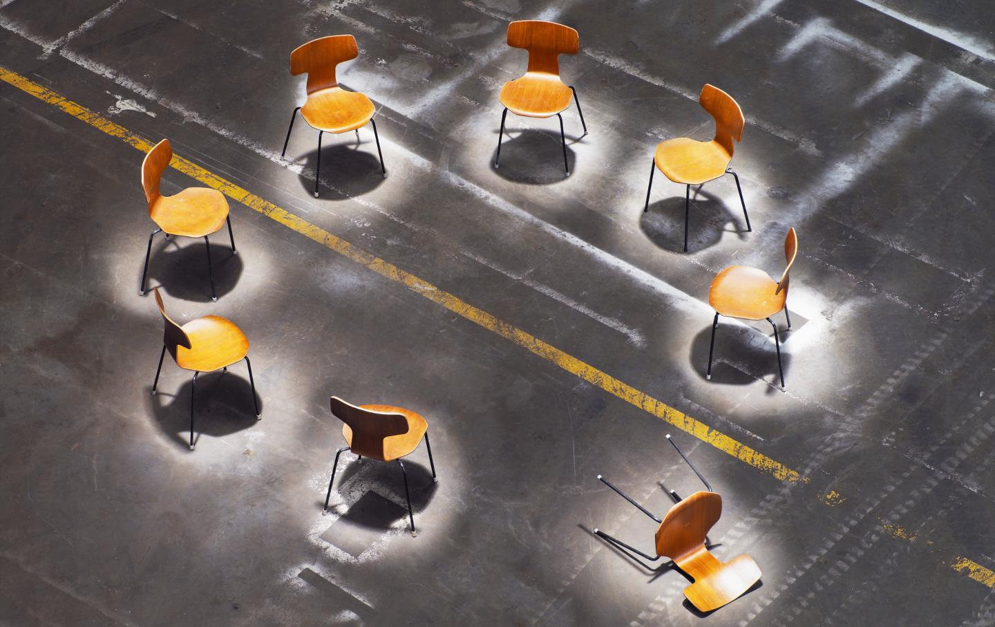 A circle of chairs with one chair tipped over, on an asphalt surface.