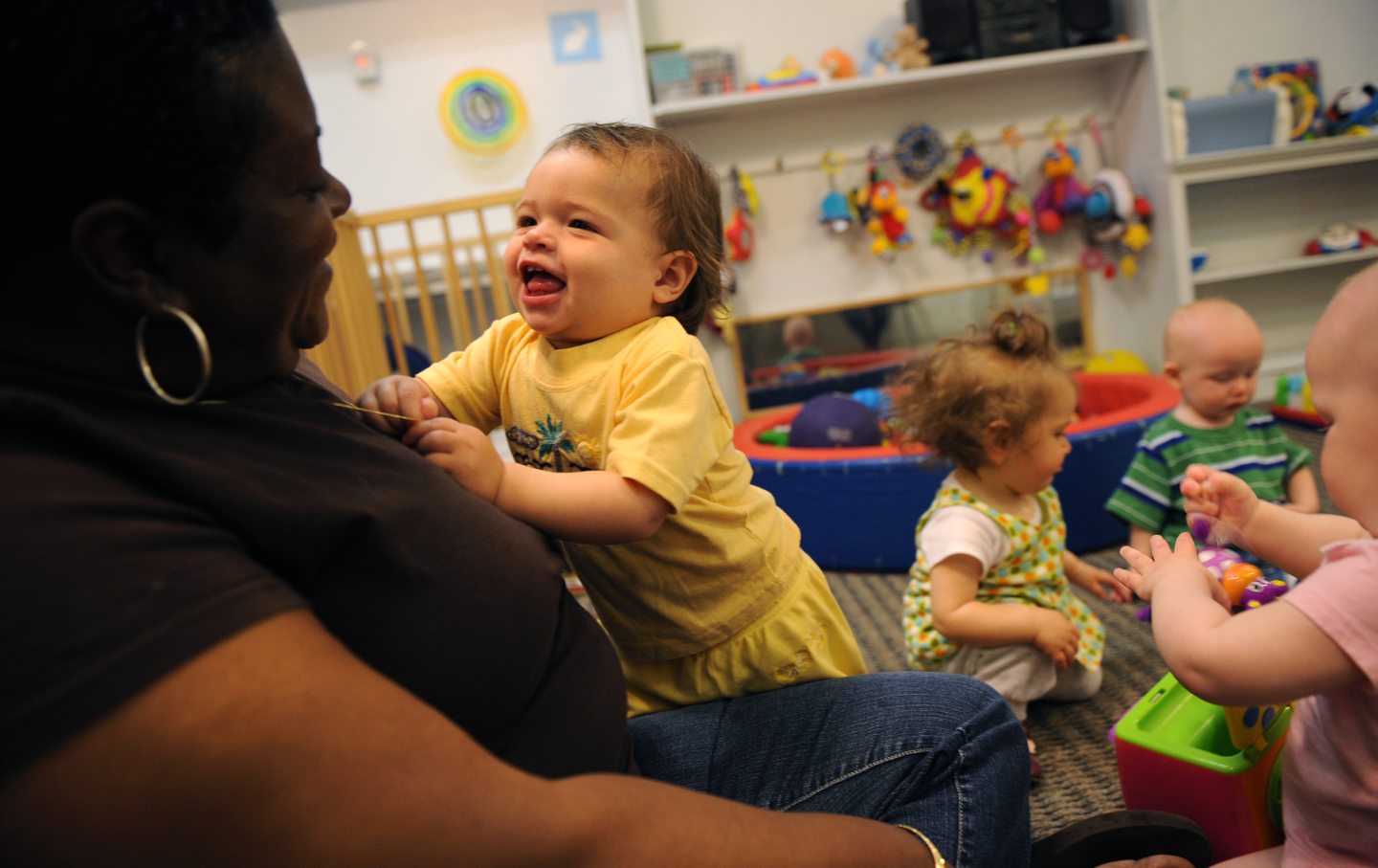 A child of about 1 year old smiles and leans on a childcare worker, with other children in the background.