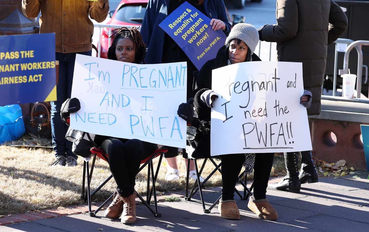 Two Black women sit on folding chairs holding signs reading 