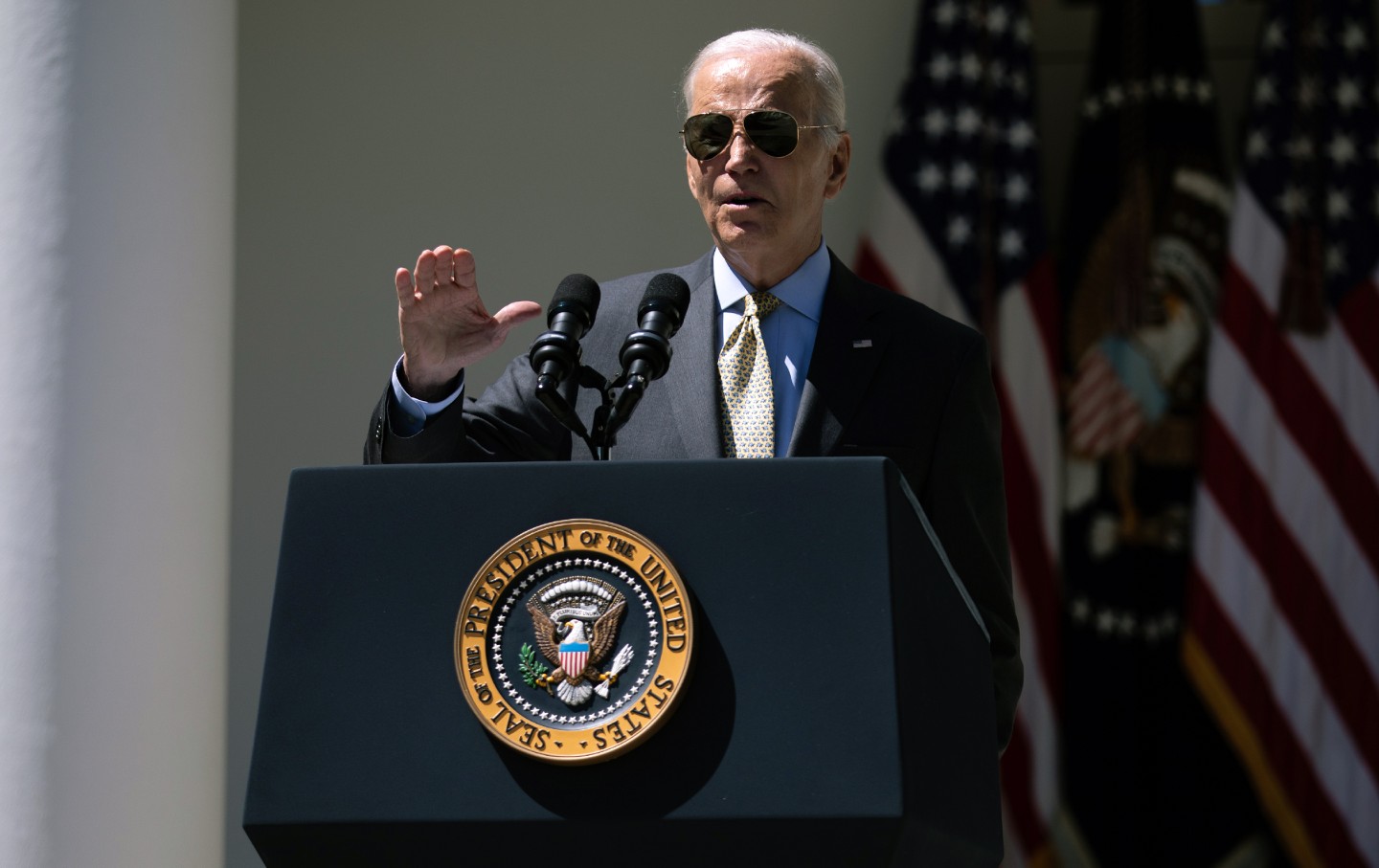 Joe Biden at an outdoor podium raising his hand and wearing sunglasses.