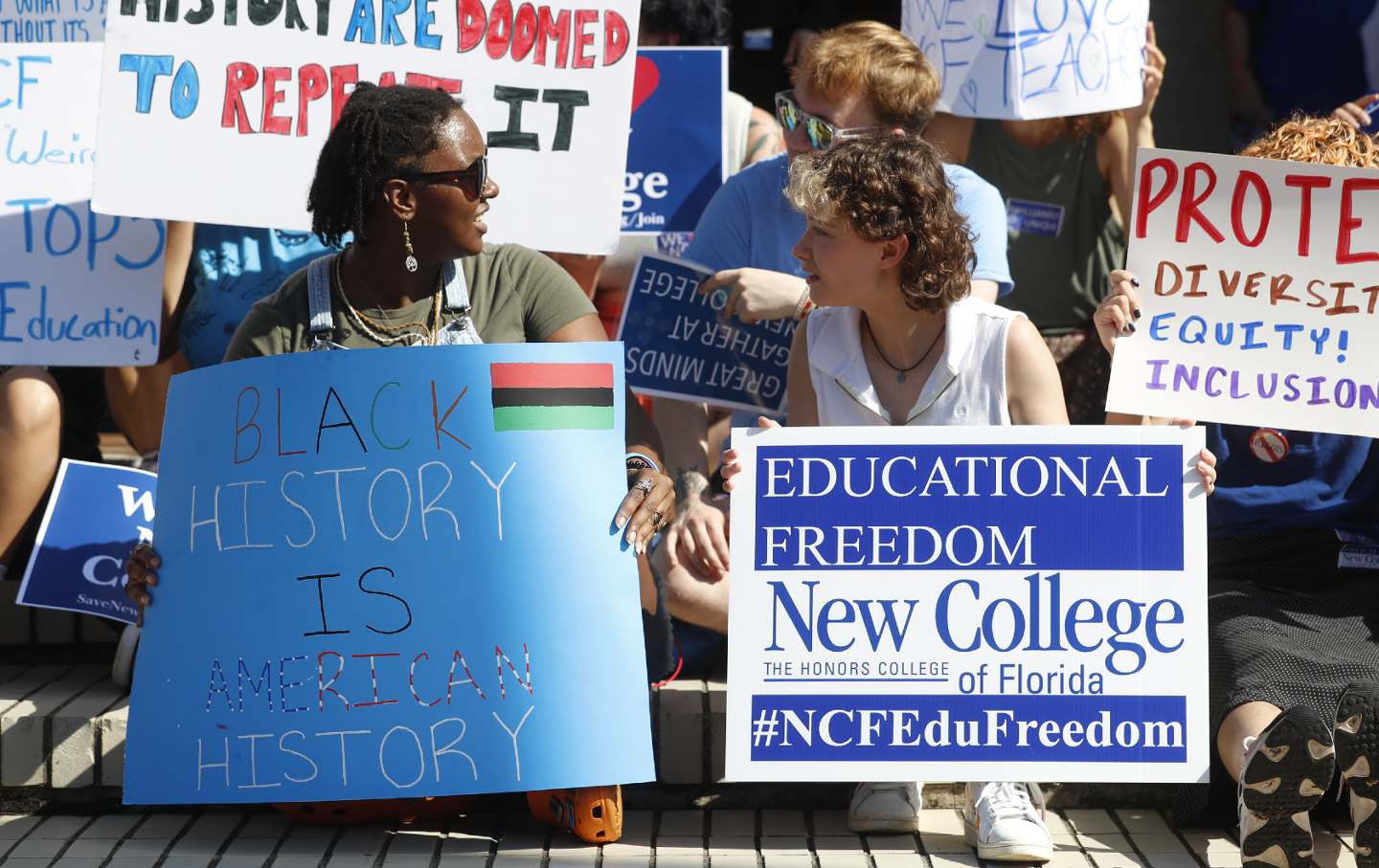 A Black student and white student speak to each other while holding signs reading, "Black History is American History" and "Educational Freedom New College."