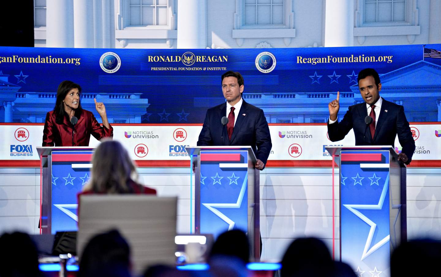 Nikki Haley, Ron DeSantis, and Vivek Ramaswamy at podiums at the Republican presidential debate. Haley and Ramaswamy have their index fingers raised, while DeSantis stares into the middle distance.
