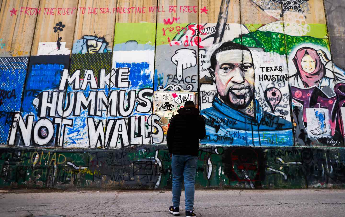 A person in jeans and a black sweatshirt looks at a brightly-colored mural depicting George Floyd as well as a sign reading "Make Hummus Not Walls."
