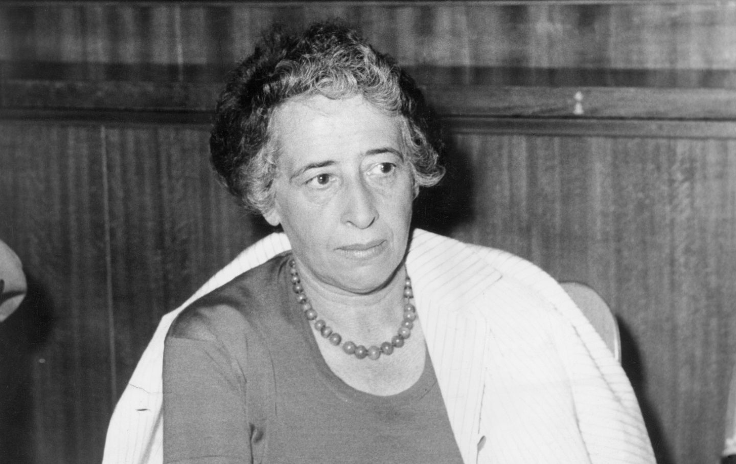 Hannah Arendt, in a black and white photograph, sits in front of a wood paneled wall wearing a white jacket