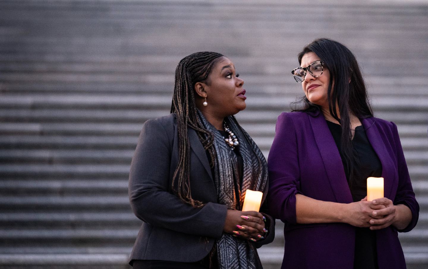 Cori Bush speaks to Rashida Tlaib while both hold candles in front of steps