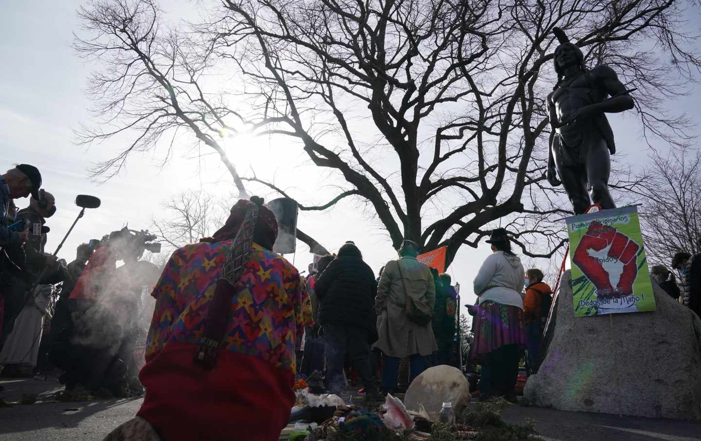 A photo of a group of people gathered, from behind, looking up at a bare tree and a statue.