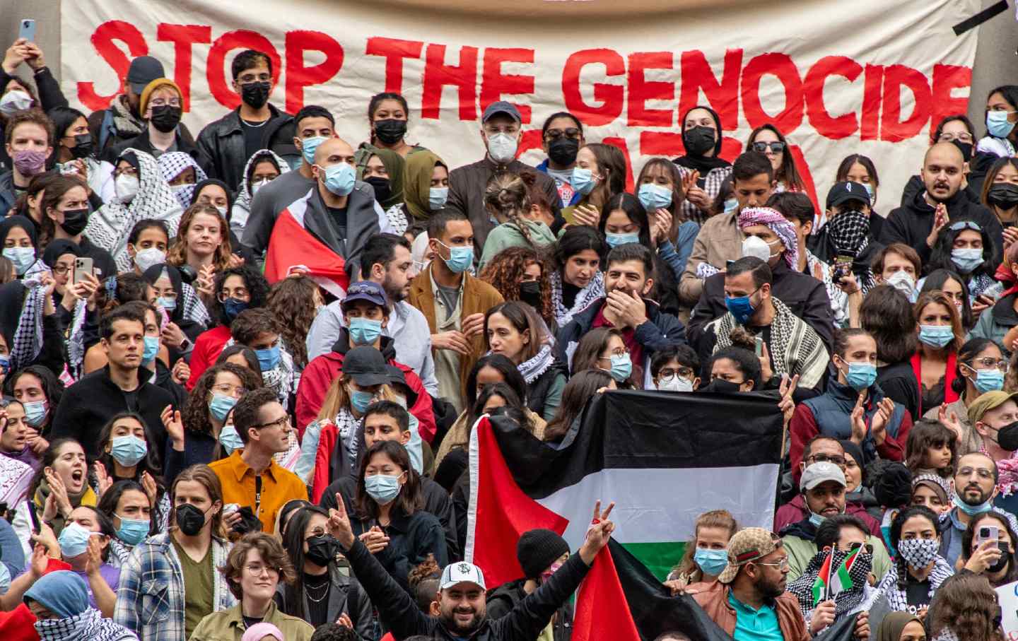A crowd of people stands in front of a banner reading "Stop the genocide"