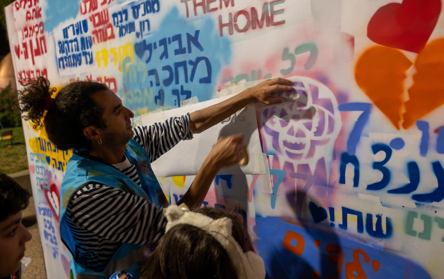 An adult with two children pastes paper onto a wall covered in graffiti