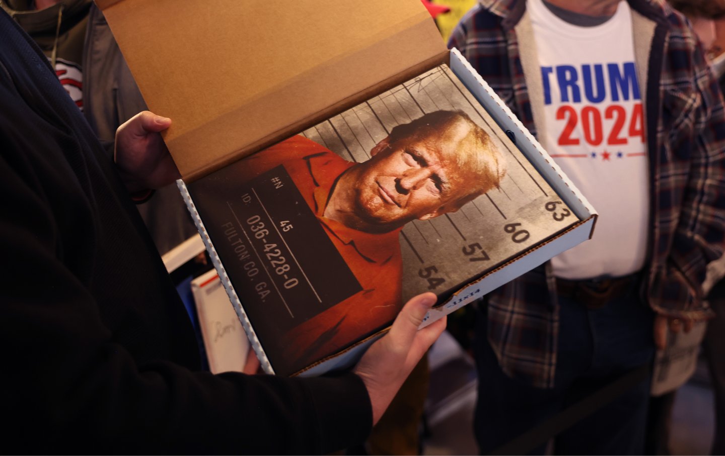 : A supporter holds a print depicting the mugshot of Republican presidential candidate former President Donald Trump as he waits for him to arrive at a commit to caucus campaign event at the Whiskey River bar on December 02, 2023 in Ankeny, Iowa.
