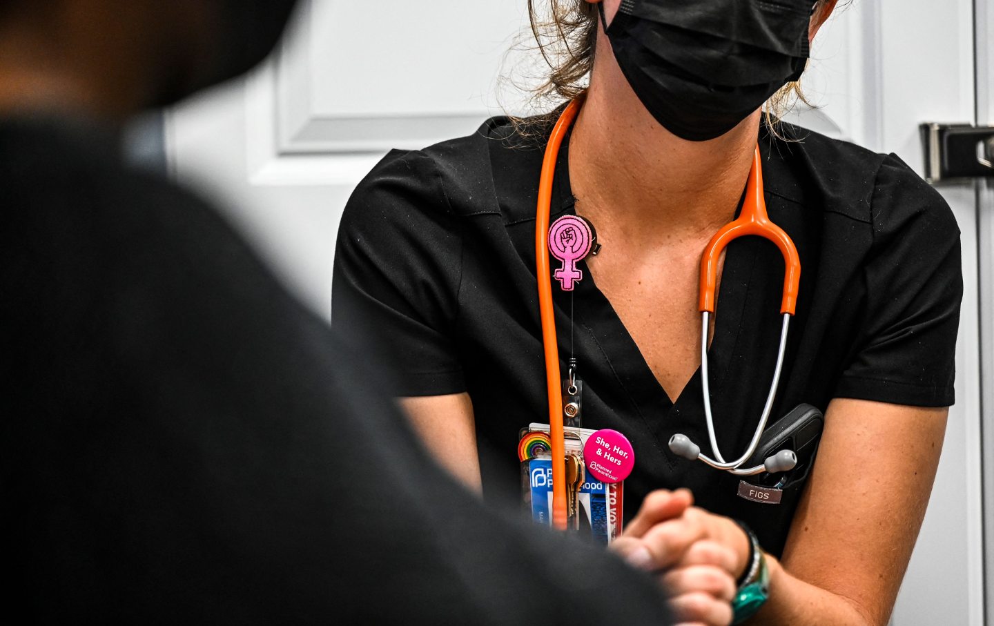A medical provider in black scrubs and a black mask, wearing a rainbow pin and woman sign pin, with a stethoscope around her neck, clasping the hands of a patient.