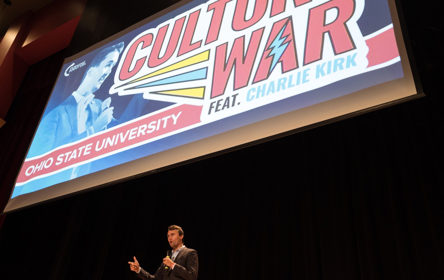 Charlie Kirk speaking on a stage under a giant banner reading 