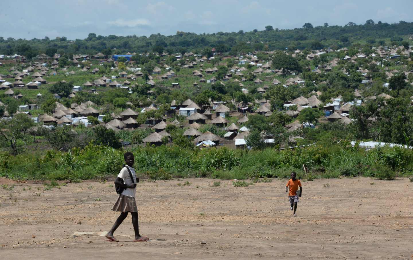 Two children walk along a dirt road in front of a field of huts.