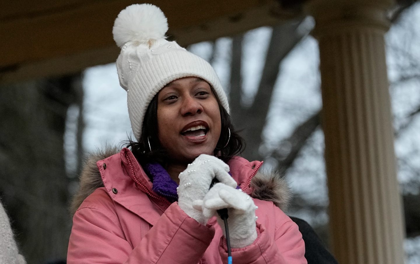 Brittany Watts, dressed in a warm pink coat and white gloves and a hat, speaking outside at a microphone.