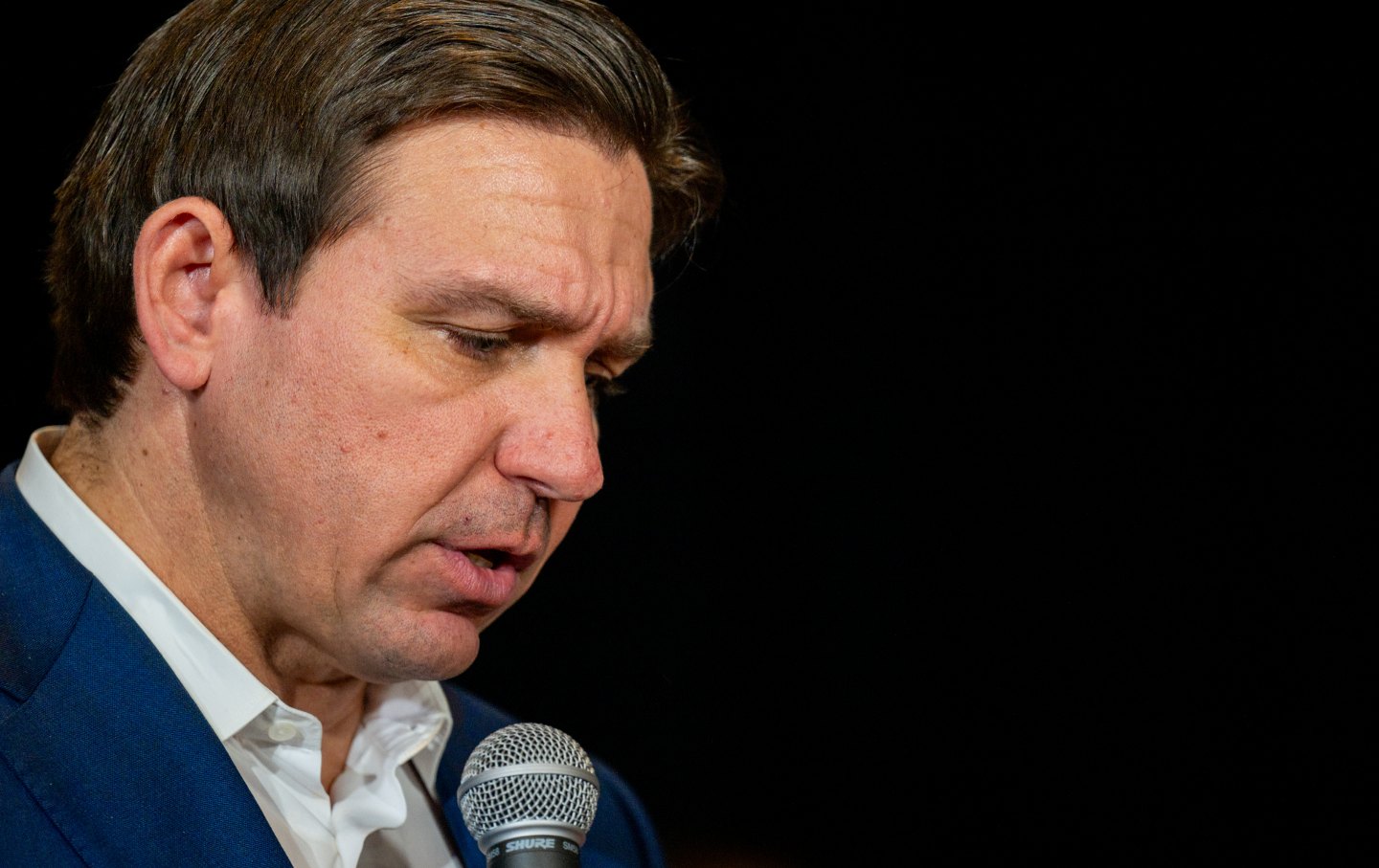 A close-cropped photo of Ron DeSantis holding a microphone, looking down.