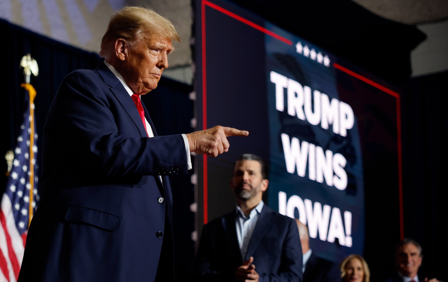 Donald Trump, on stage, points in front of a large sign reading 