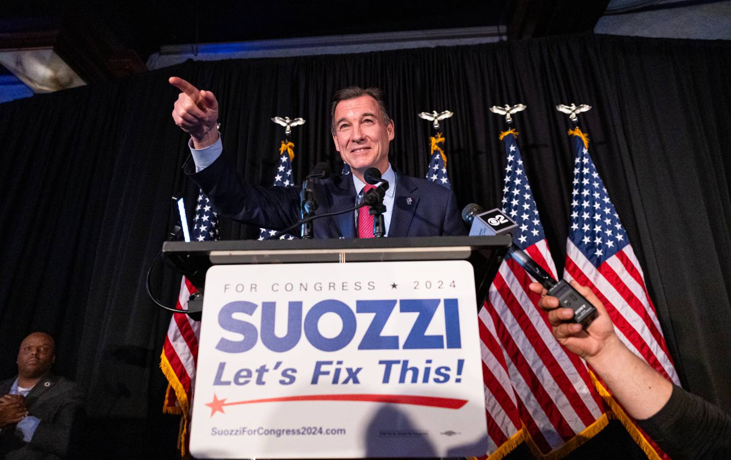Tom Suozzi at the stage on election night, smiling in front of American flags and a sign reading 