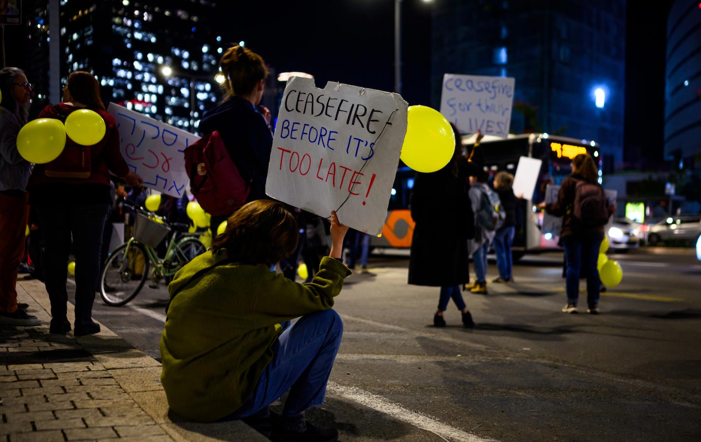 A protester sits on the sidewalk holding a sign that reads, “CEASEFIRE BEFORE IT’S TOO LATE” outside the main entrance to The Kirya on March 4, 2024, in central Tel Aviv, Israel.