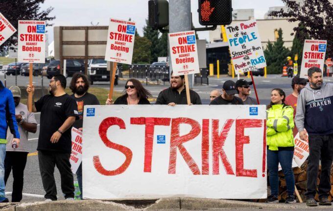 Workers holding picket signs stand in the street, with a banner reading STRIKE in red.