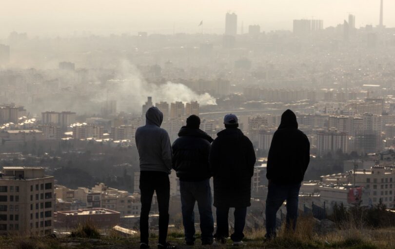 Men watch from a hillside as a plume of smoke rises after an explosion on March 2, 2026, in Tehran, Iran.