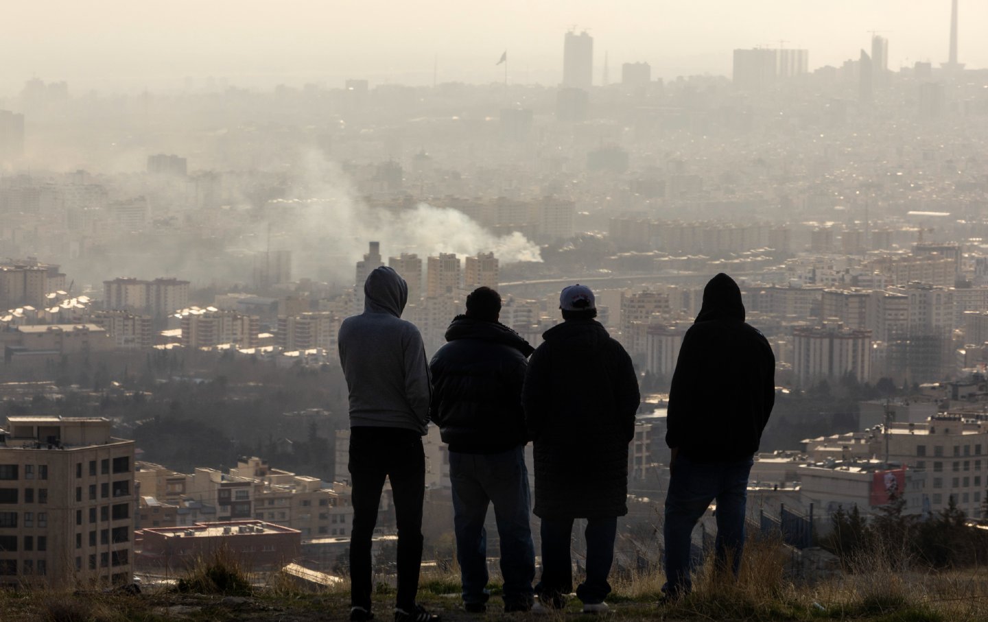 Men watch from a hillside as a plume of smoke rises after an explosion on March 2, 2026, in Tehran, Iran.